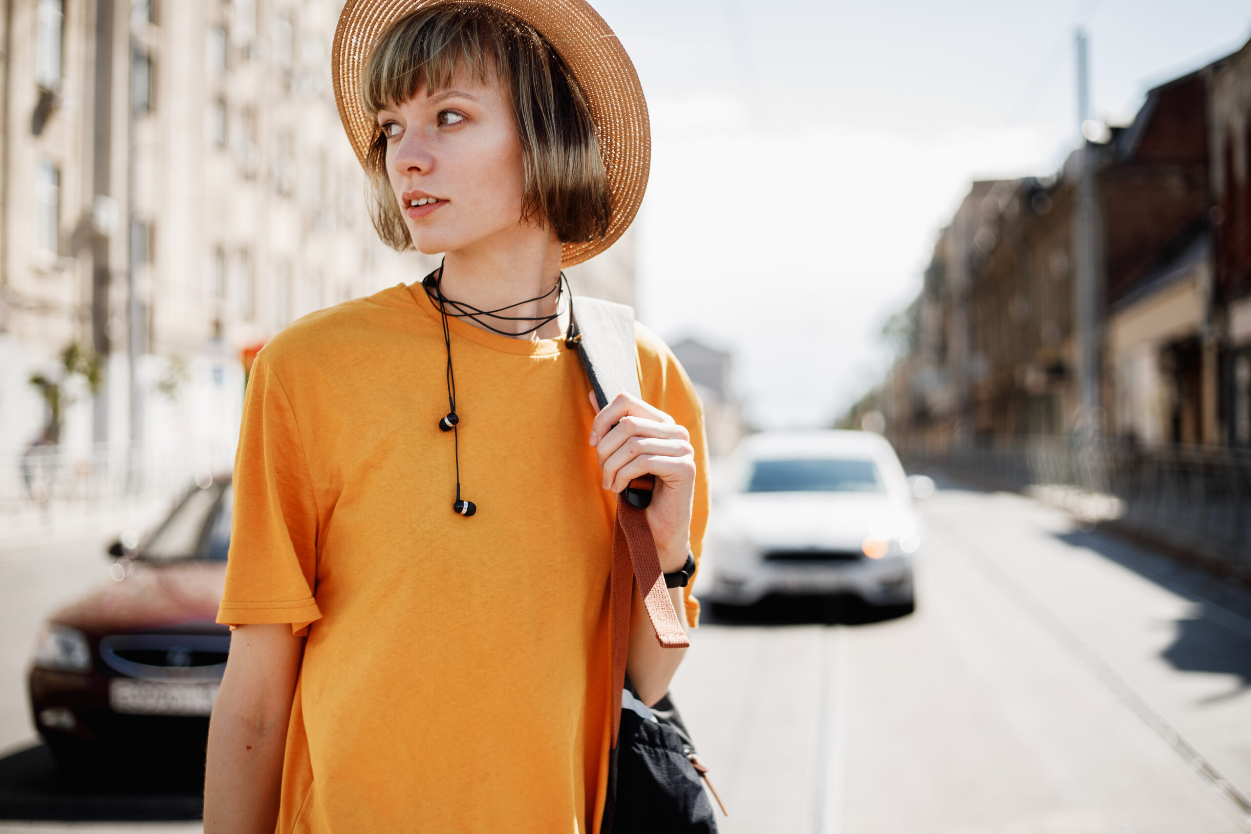 Young girl with headphones in a yellow T-shirt and a straw hat walks with a backpack along a city street on a summer sunny day .